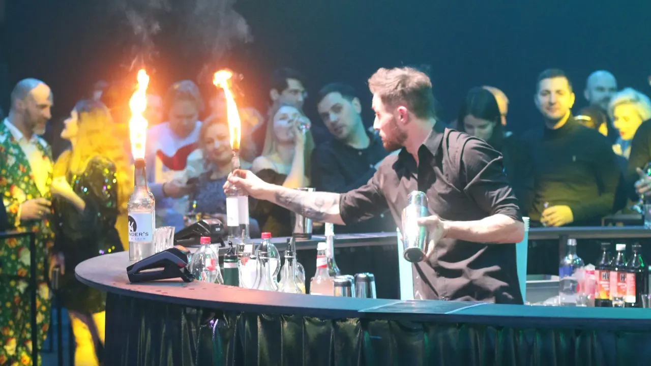 A show bartender behind the stage bar holding flaming bottles, the crowd behind him